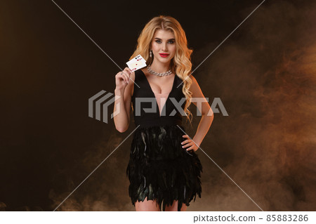 Blonde female in black dress with feathers and necklace. She is smiling, showing two aces, posing on black smoky background. Poker, casino. Close-up 85883286