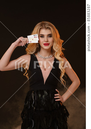Blonde woman in black dress with feathers and necklace. Smiling, showing two playing cards, posing on black smoky background. Poker, casino. Close-up 85883287