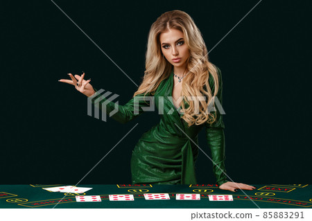 Girl in stylish dress is showing something, leaning on playing table with cards on it, posing on green background. Poker, casino. Close-up, copy space Girl in stylish dress is showing something, leaning on playing table with cards on it, posing on green background. Poker, casino. Close-up, copy space 85883291