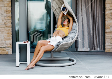 A young woman is resting in a suspended swing on the terrace of the house with a magazine in her hands 85884187
