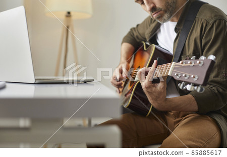 Musical performance online. Man plays acoustic guitar at home in front of online audience or provides music lesson using laptop. Cropped image of serious man with guitar in his hands. Selective focus. Musical performance online. Man plays acoustic guitar at home in front of online audience or provides music lesson using laptop. Cropped image of serious man with guitar in his hands. Selective focus. 85885617