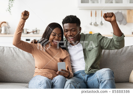 Happy black lovers using smartphone together and gesturing, home interior Happy black lovers using smartphone together and gesturing, home interior 85888386