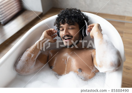 Handsome indian man wearing wireless headphones and singing while lying in hot bubbly bath at home 85888490