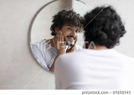 Attractive bearded indian man touching face applying moisturizer on face, standing near mirror in modern bathroom Attractive bearded indian man touching face applying moisturizer on face, standing near mirror in modern bathroom 85888543
