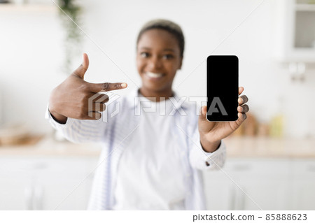Cheerful young black woman shows on smartphone with blank screen at scandinavian kitchen interior, selective focus 85888623