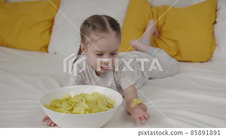 little child eats crispy fries lying on the bed, kid bed food, delicious baby food chips, snacks, happy family, children's life at home, baby girl gnaws fried vegetable, not healthy human food. little child eats crispy fries lying on the bed, kid bed food, delicious baby food chips, snacks, happy family, children's life at home, baby girl gnaws fried vegetable, not healthy human food. 85891891
