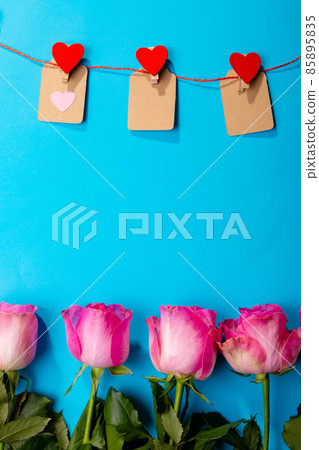 Red heart shapes on clothespins hanging from clothesline over pink roses against blue background Red heart shapes on clothespins hanging from clothesline over pink roses against blue background 85895835