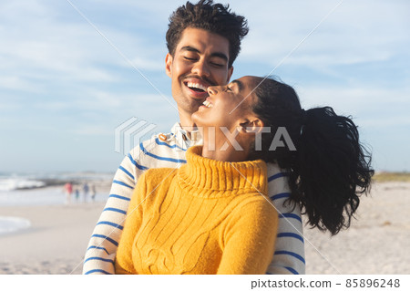 Happy biracial man embracing cheerful young girlfriend from behind at beach against sky Happy biracial man embracing cheerful young girlfriend from behind at beach against sky 85896248