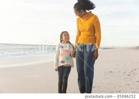 Happy biracial mother and daughter holding hands while looking at each other on beach against sky Happy biracial mother and daughter holding hands while looking at each other on beach against sky 85896252