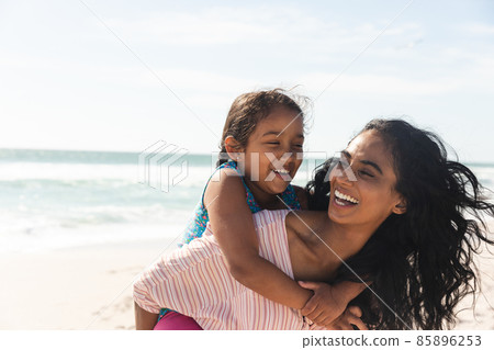 Cheerful biracial woman giving piggyback ride to daughter at beach against sky with copy space Cheerful biracial woman giving piggyback ride to daughter at beach against sky with copy space 85896253