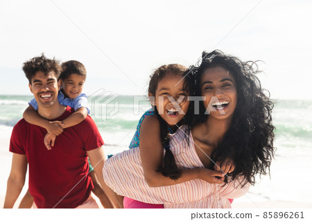 Portrait of happy multiracial parents giving piggyback rides to kids at beach with copy space Portrait of happy multiracial parents giving piggyback rides to kids at beach with copy space 85896261