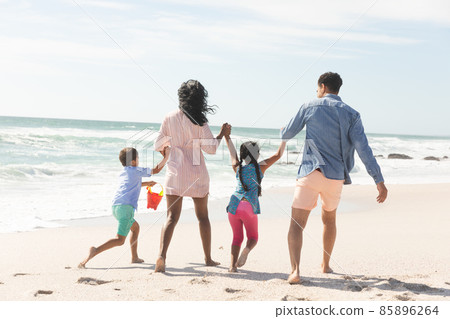 Full length rear view of playful multiracial family holding hands walking at beach during sunny day Full length rear view of playful multiracial family holding hands walking at beach during sunny day 85896264