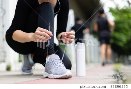 Crop unrecognizable woman runner wearing hygiene protective face mask sitting and tieing shoe rope during workout running in city park. Concept of healthcare in time of coronavirus outbreak 85896355