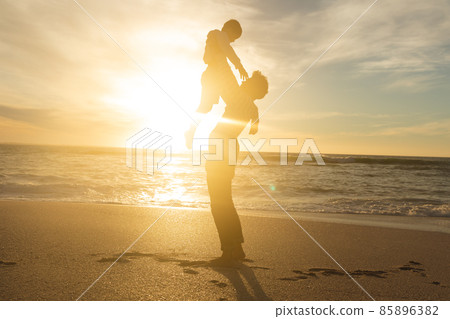 Side view of back lit biracial father holding aloft son on shore at beach against sky during sunset Side view of back lit biracial father holding aloft son on shore at beach against sky during sunset 85896382