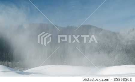Artificial snow falls on background of spruce forest in mountains at ski resort. White snow-covered mountain slope for skiing on sunny frosty day in winter. Working snow cannon, snowgun on blue sky Artificial snow falls on background of spruce forest in mountains at ski resort. White snow-covered mountain slope for skiing on sunny frosty day in winter. Working snow cannon, snowgun on blue sky 85898568