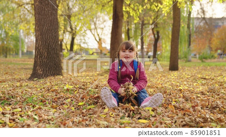 happy girl throws autumn yellow leaves up and laughs, kid is having fun playing in city nature park outdoors, little child with school backpack sits under leaf fall, first grader school yard having happy girl throws autumn yellow leaves up and laughs, kid is having fun playing in city nature park outdoors, little child with school backpack sits under leaf fall, first grader school yard having 85901851