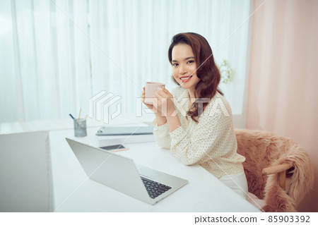 Asian female relaxing sitting in front of laptop, drinking from mug taking a break Asian female relaxing sitting in front of laptop, drinking from mug taking a break 85903392