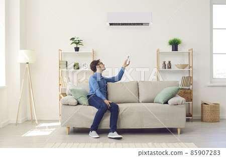 Young man turns on air conditioner with remote control while relaxing on couch at home. Man sits under air conditioning system and makes comfortable temperature for himself. Home comfort management. 85907283
