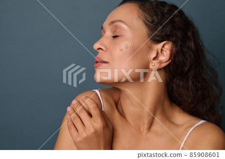 Charming dark-skinned beautiful woman with closed eyes posing on a gray background with dots of liquid foundation on her face. Perfect natural makeup and skin care concept. Copy space for advertising 85908601
