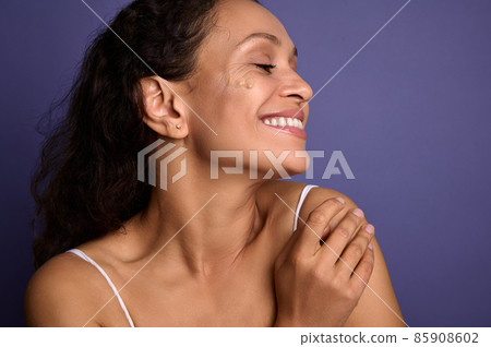 Beautiful dark-haired African woman with clean healthy glowing skin posing with spots of liquid tonal foundation on her face, smiles toothy smile looking aside over purple background with copy space. 85908602