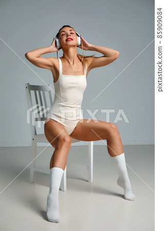 Banner of young happy girl in white swimsuit listening to music in white chair, wearing wireless headhones, isolated on gray background with copy space in studio 85909084