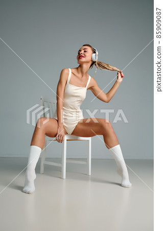 Banner of young happy girl in white swimsuit listening to music in white chair, wearing wireless headhones, isolated on gray background with copy space in studio 85909087