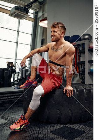 Strength concept. Male athlete using hammer during fitness workout. Fitness man beating rubber tire in gym. Muscular sportsman performing intensity training with sledgehammer in sport club 85909187