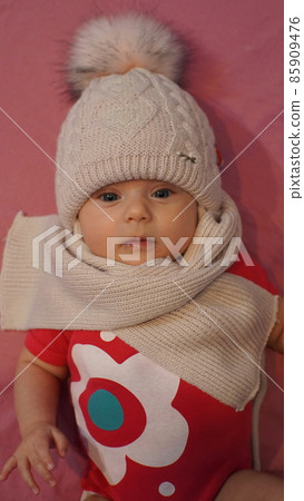 A beautiful, cute baby with blue eyes in a hat with a bubo, large knit, acrylic, handmade and a scarf looks at the camera on a pink background. In a red t-shirt. Dressed before going out in winter 85909476