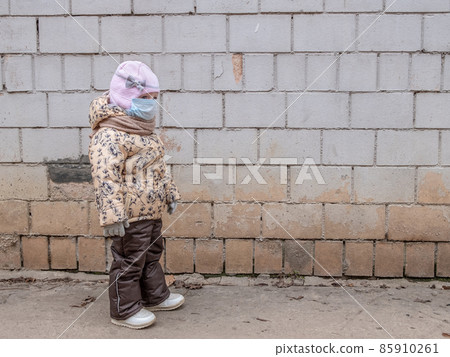 A little child girl in a protective mask on her face stands near an old wall.  85910261