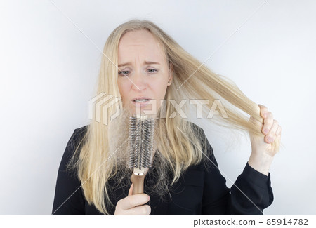 Close-up of a girl whose hair is falling out. There is a large tuft of tangled curls on the comb. The blonde looks upset into the frame. The concept of women problems with the scalp 85914782