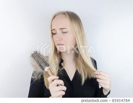 Close-up of a girl whose hair is falling out. There is a large tuft of tangled curls on the comb. The blonde looks upset into the frame. The concept of women problems with the scalp 85914784