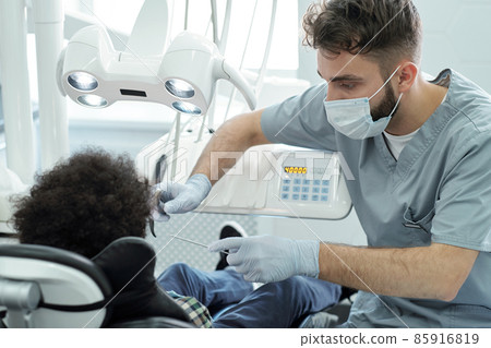 Dentist in uniform, mask and gloves holding dental drill and instrument by mouth of little patient Dentist in uniform, mask and gloves holding dental drill and instrument by mouth of little patient 85916819