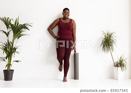 Joyful chubby black lady standing by wall with fitness mat 85917345