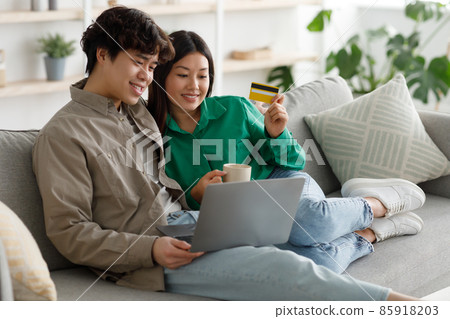 Happy Asian couple shopping online via laptop and credit card, ordering things in internet, sitting on sofa at home Happy Asian couple shopping online via laptop and credit card, ordering things in internet, sitting on sofa at home 85918203