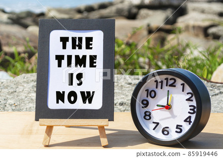 Motivational quote on standing notepad with black alarm clock. The time is now Motivational quote on standing notepad with black alarm clock. The time is now 85919446