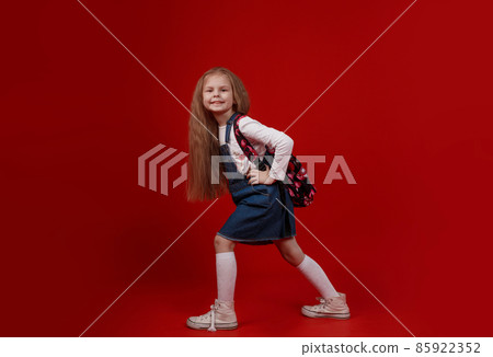 a beautiful little schoolgirl in a denim dress with a backpack on her shoulders walks on a red isolated background. 85922352