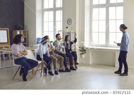 Leader of the startup team listens to the suggestions of his colleagues at a meeting in the office. Woman surrounded by her colleagues sitting on chairs raises her hand wanting to talk about new ideas Leader of the startup team listens to the suggestions of his colleagues at a meeting in the office. Woman surrounded by her colleagues sitting on chairs raises her hand wanting to talk about new ideas 85922522