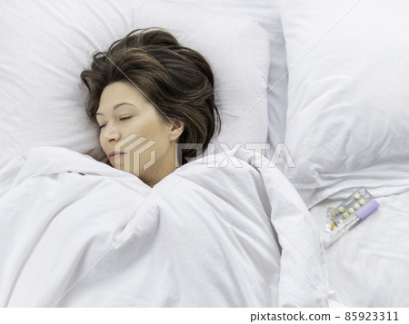Top view on sick woman lying in bed. Thermometer and pills on white bed linen. Morning at home. Top view on sick woman lying in bed. Thermometer and pills on white bed linen. Morning at home. 85923311
