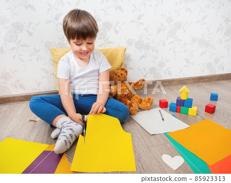 Toddler boy learns to cut colored paper with scissors. Kid sits on floor in kids room with toy blocks and teddy bear. Educational classes for children. Developing feeling sensations and fine motor Toddler boy learns to cut colored paper with scissors. Kid sits on floor in kids room with toy blocks and teddy bear. Educational classes for children. Developing feeling sensations and fine motor 85923313