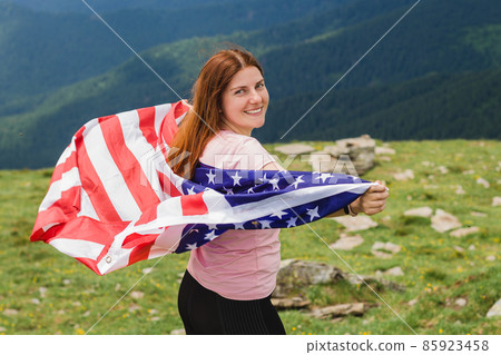 Girl with American Flag looking out at landscape. Young woman stands in the mountains, holding the US flag in her arms high. Flag fluttering in the wind 85923458