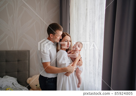 a happy family with a newborn baby by the window. mom, dad and baby. a happy family with a newborn baby by the window. mom, dad and baby. 85924805