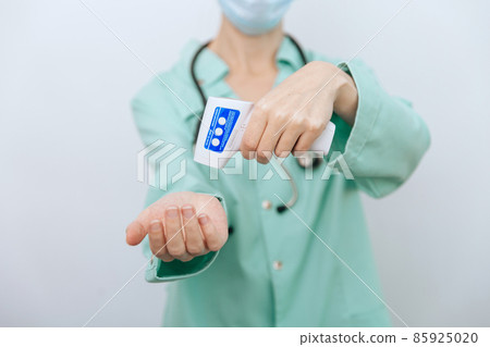 Temperature measurement gun in doctor hands. Close-up shot of doctor wearing protective surgical mask ready to use infrared isometric thermometer gun to check body temperature for virus symptoms. Temperature measurement gun in doctor hands. Close-up shot of doctor wearing protective surgical mask ready to use infrared isometric thermometer gun to check body temperature for virus symptoms. 85925020
