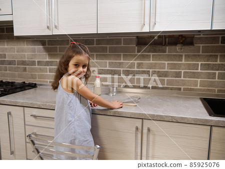 Adorable Caucasian baby girl standing on a chair by kitcheb countertop and smiles looking at camera through her shoulders 85925076