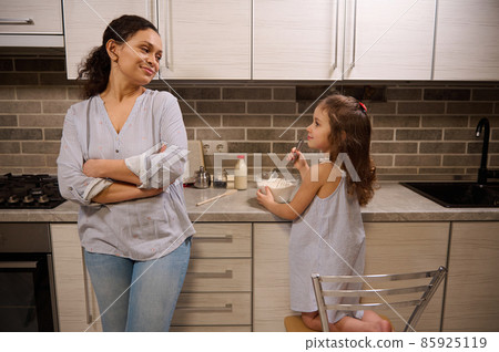 Beautiful woman, happy loving mother leaning at kitchen countertop and admiring her adorable daughter cute baby girl mixing ingredients in a glass bowl while learning kneading pancakes dough 85925119