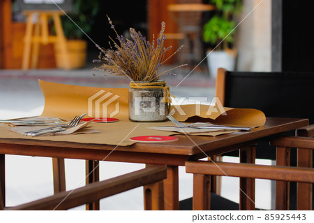 Wooden dining table, chairs on a street cafe terrace. Cutlery is on table, a vase with a flower arrangement, lavender stands nearby. Cozy restaurant table without visitors in European. Rustic serving. Wooden dining table, chairs on a street cafe terrace. Cutlery is on table, a vase with a flower arrangement, lavender stands nearby. Cozy restaurant table without visitors in European. Rustic serving. 85925443