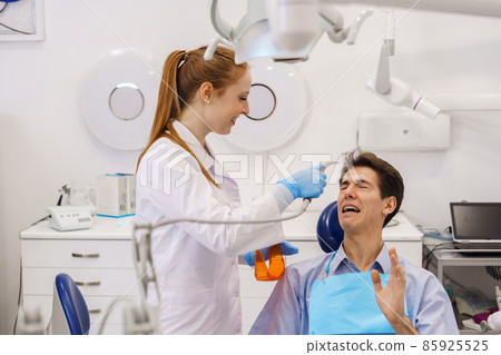 Happy female doctor with ginger ponytail smiling and preparing to heal terrified man in modern dentist office 85925525