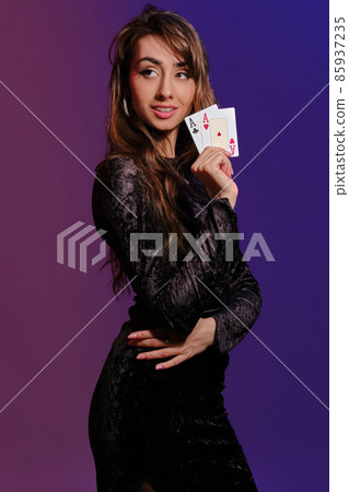 Brunette girl in black velvet dress showing two playing cards, posing sideways on coloful background. Gambling entertainment, poker, casino. Close-up. Brunette girl in black velvet dress showing two playing cards, posing sideways on coloful background. Gambling entertainment, poker, casino. Close-up. 85937235
