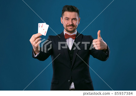 Man in black classic suit and red bow-tie showing two playing cards while posing against blue studio background. Gambling, poker, casino. Close-up. 85937384
