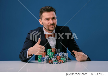 Man in black suit is sitting at white table with colored stacks of chips on it, posing on blue studio background. Gambling, poker, casino. Close-up. Man in black suit is sitting at white table with colored stacks of chips on it, posing on blue studio background. Gambling, poker, casino. Close-up. 85937439
