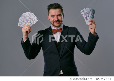 Man in black suit and red bow-tie is showing two fans of hundred dollar bills, posing on gray studio background. Gambling, poker, casino. Close-up. Man in black suit and red bow-tie is showing two fans of hundred dollar bills, posing on gray studio background. Gambling, poker, casino. Close-up. 85937485